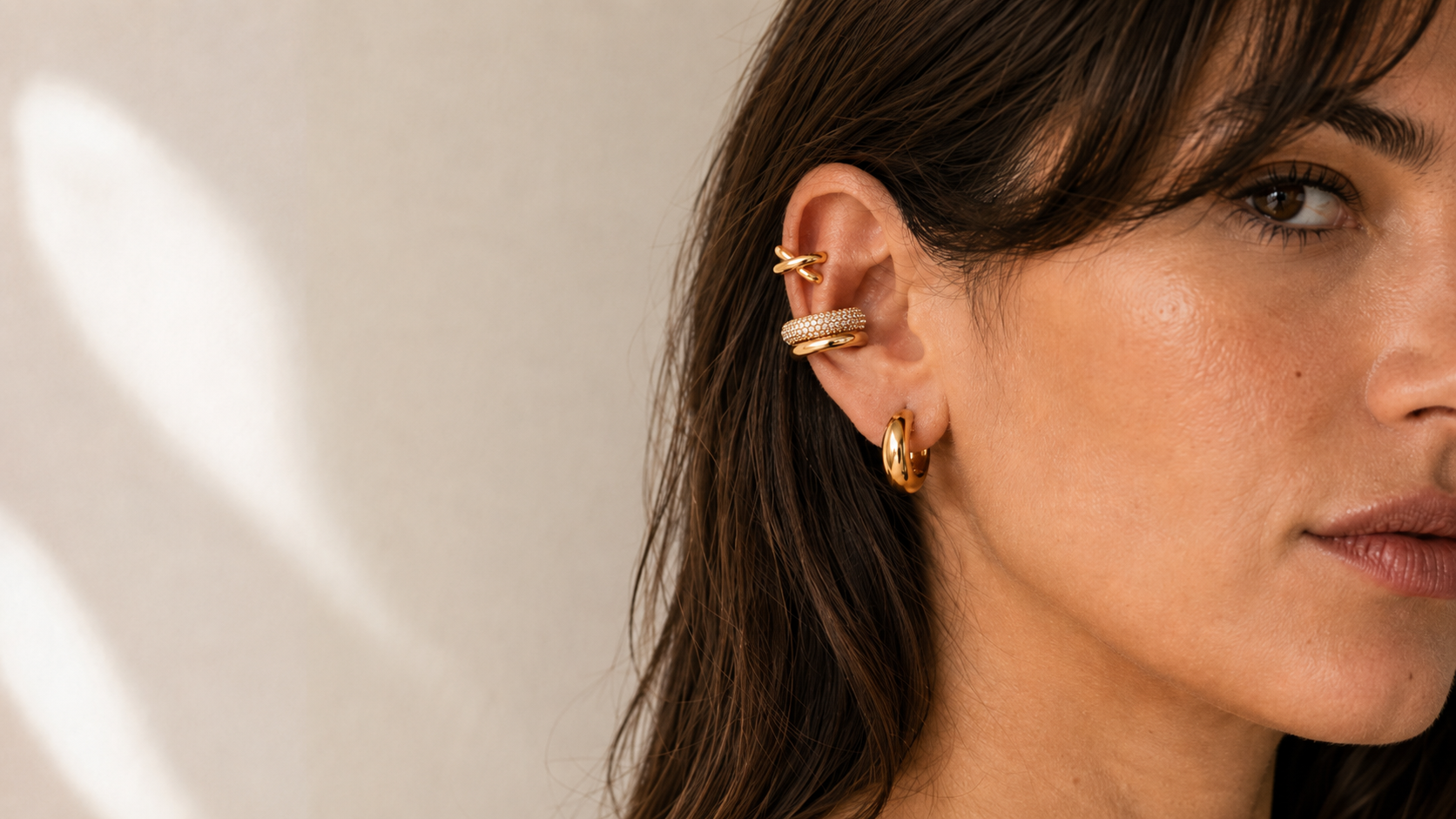 Woman's ear with three gold ear cuffs stacked on the upper ear and lobe, warm natural light, off-white background. No piercing needed. Stainless steel with PVD gold.