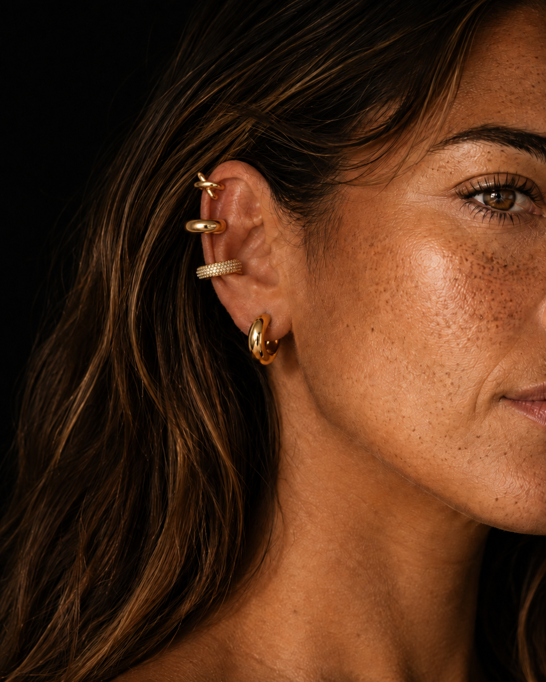 Woman's ear in profile wearing four gold ear cuffs arranged as a full stack, dark background with warm side light. No piercing needed. Stack and save up to 25%.