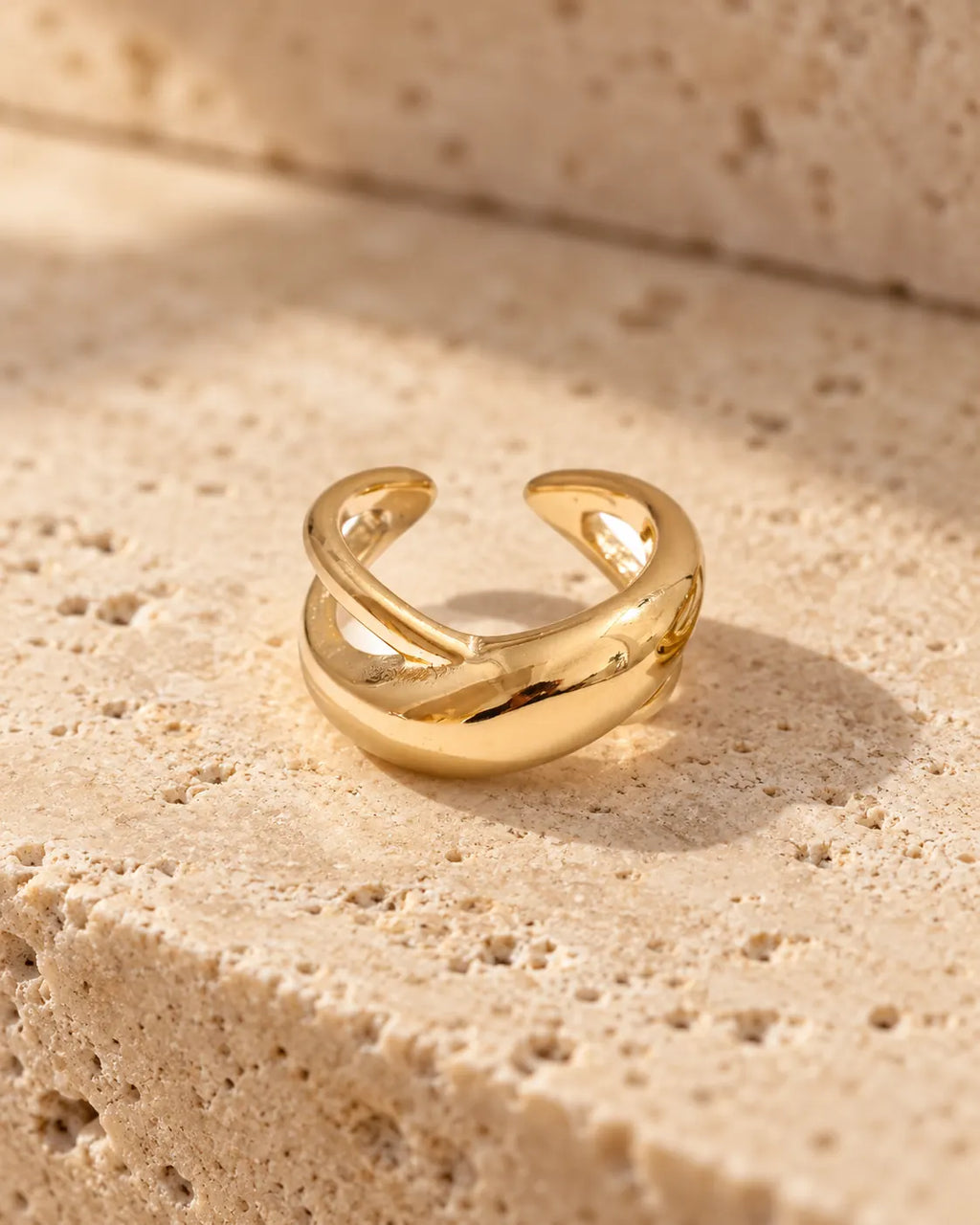 Gold Axis crossed ear cuff on travertine stone, the X-shaped bands catching warm daylight. PVD real gold plated stainless steel.