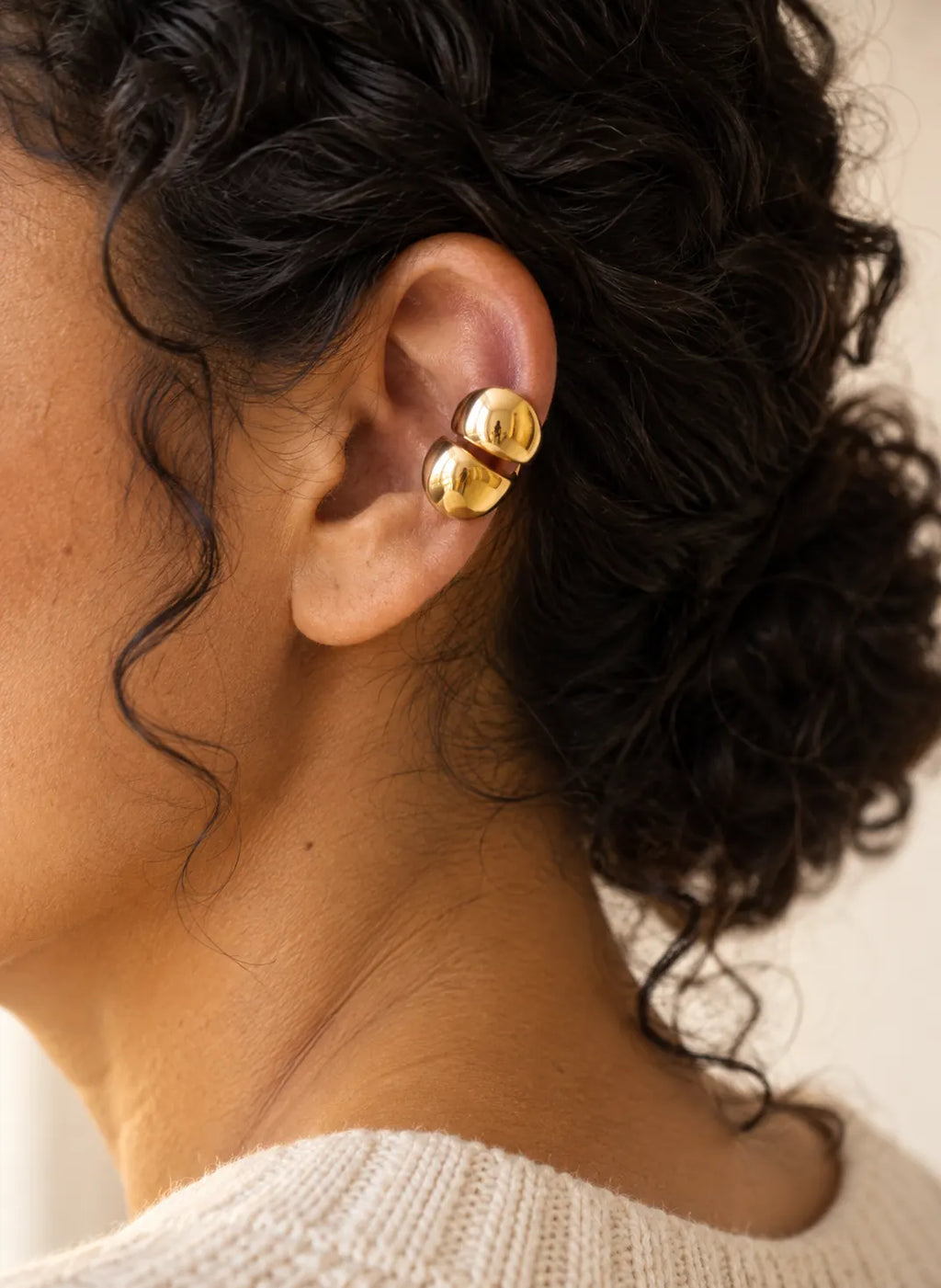 Gold double ear cuff seen from behind on a woman's ear, curly hair pulled back in a low bun. No piercing needed.