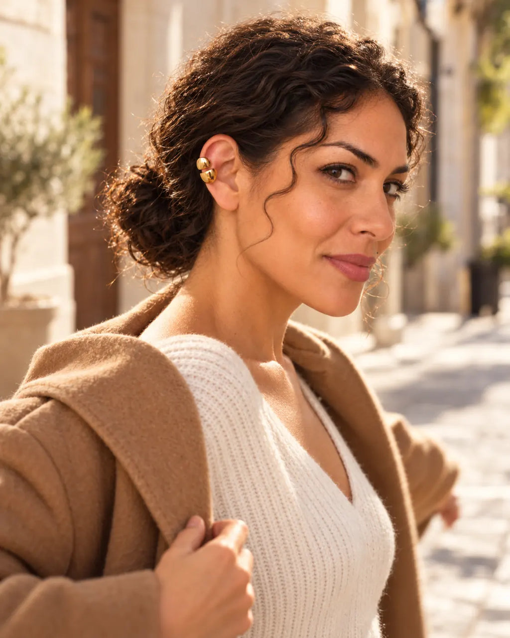Woman pulling on a camel coat on a sunny street, wearing a gold double ear cuff on her upper ear. No piercing needed.