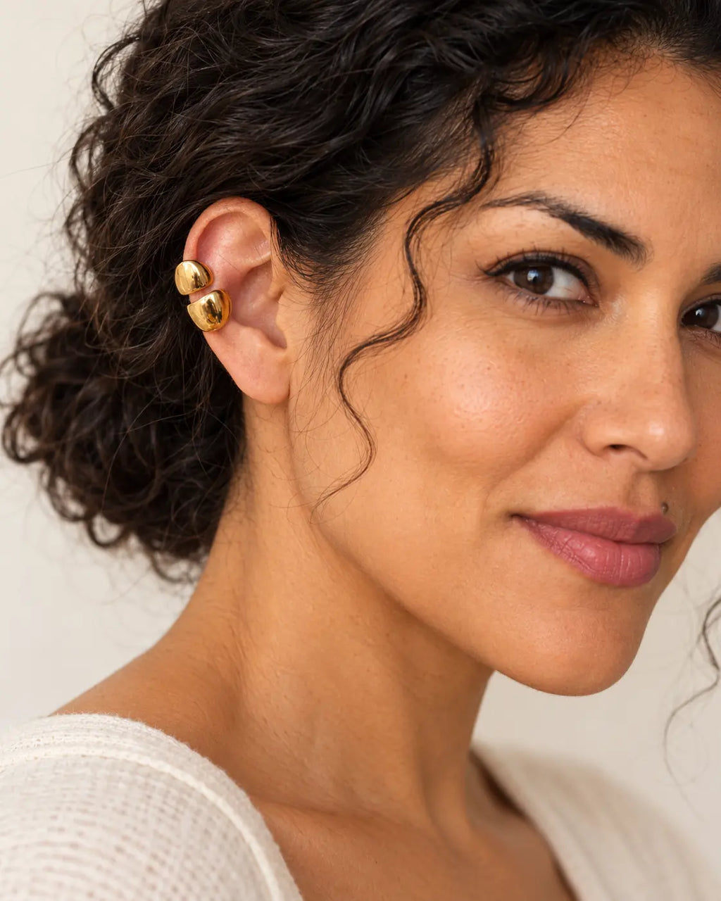 Gold double ear cuff on a woman's upper ear in three-quarter view, warm neutral background. No piercing needed.