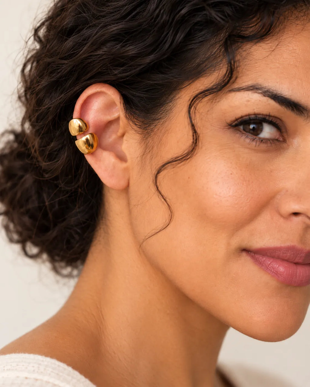 Gold double ear cuff with two polished domes on a woman's upper ear, curly hair pulled back, warm neutral background. No piercing needed.
