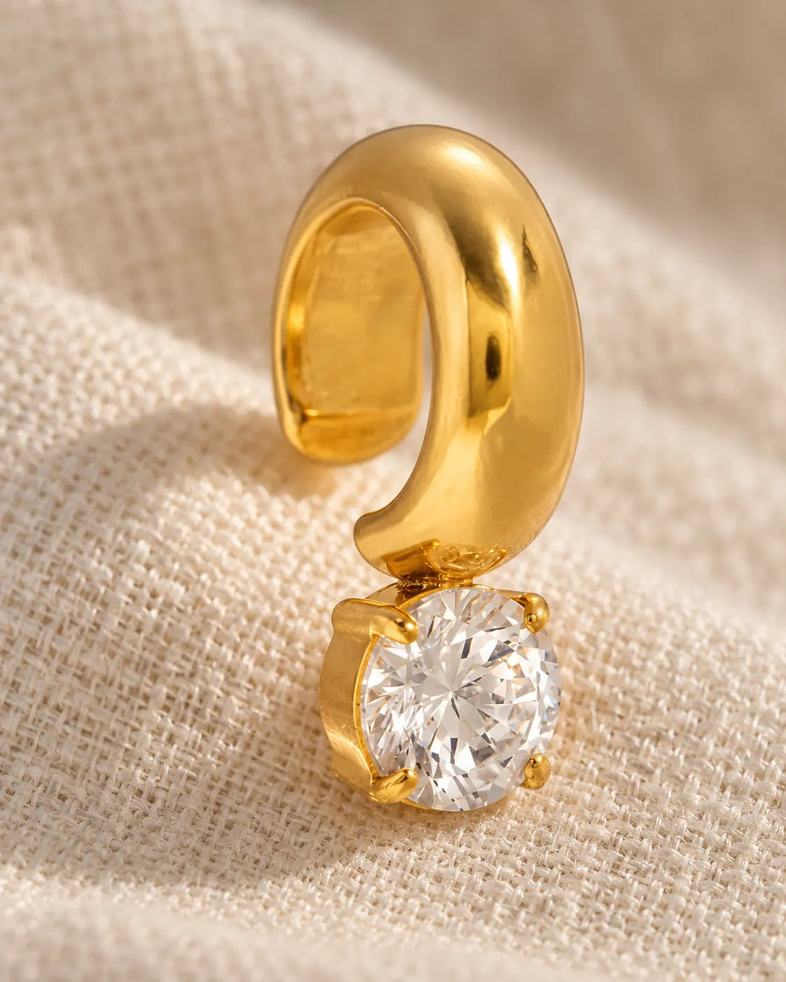 Close-up of the gold Glint Drop Ear Cuff on cream linen: the four-claw setting holding one round stone below the domed band.
