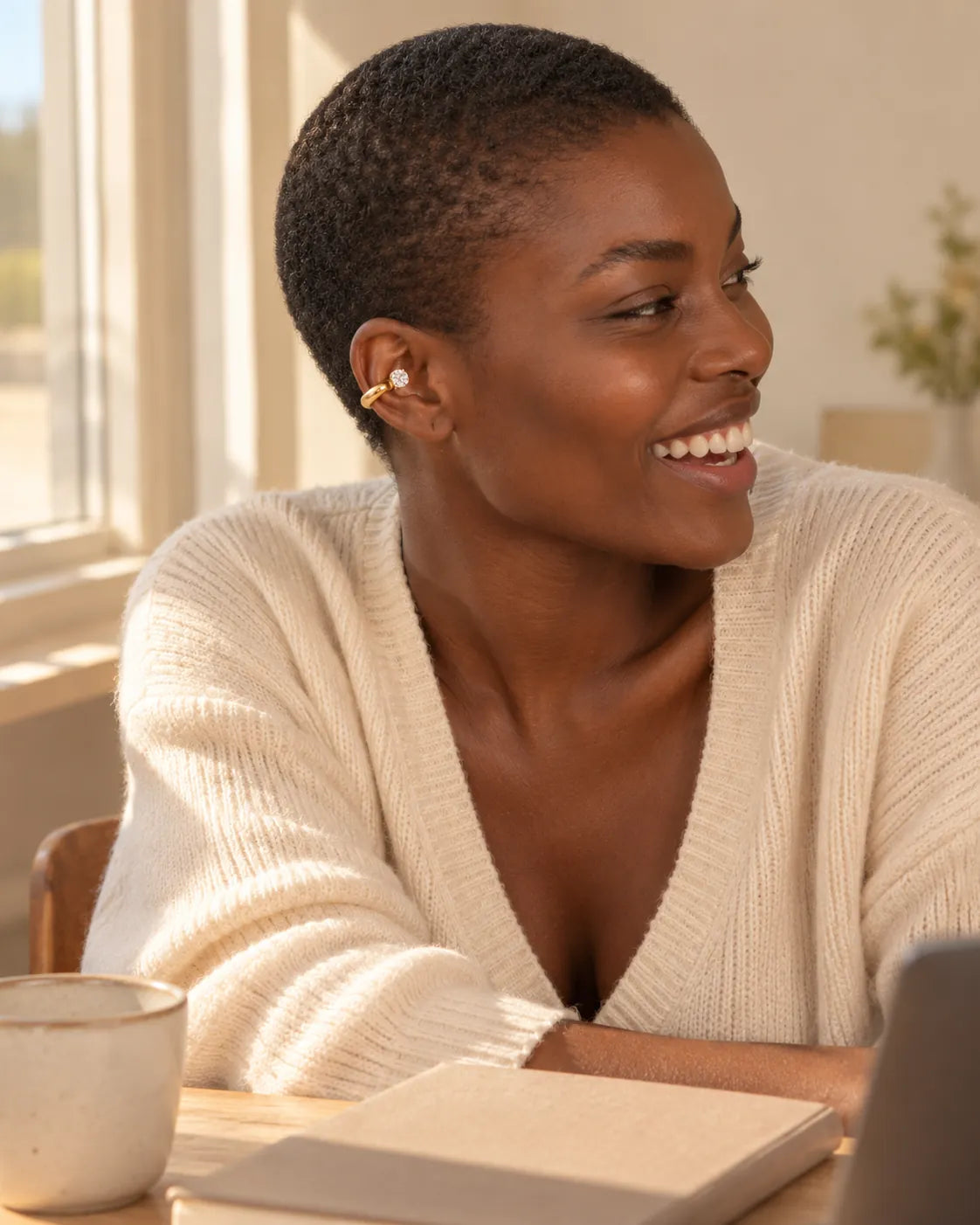 Woman laughing at a light wooden desk by a window, wearing a gold drop ear cuff on her upper ear. No piercing needed.