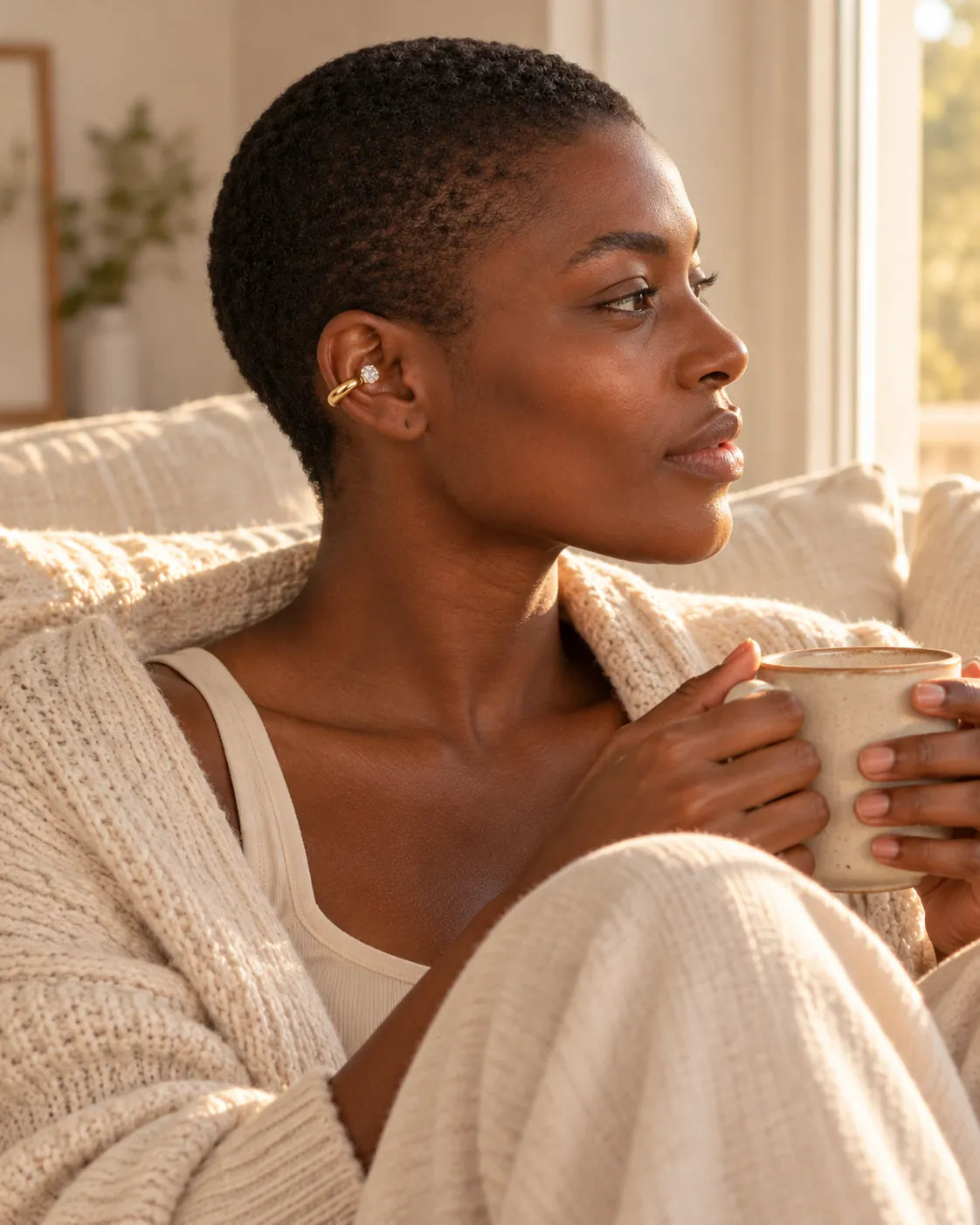 Woman on a linen sofa holding a mug and looking toward the window, wearing a gold drop ear cuff on her upper ear. No piercing needed.