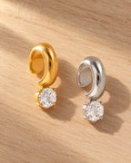 Glint Drop Ear Cuff in gold and silver on light oak wood: a chunky polished band with one round stone hanging below.