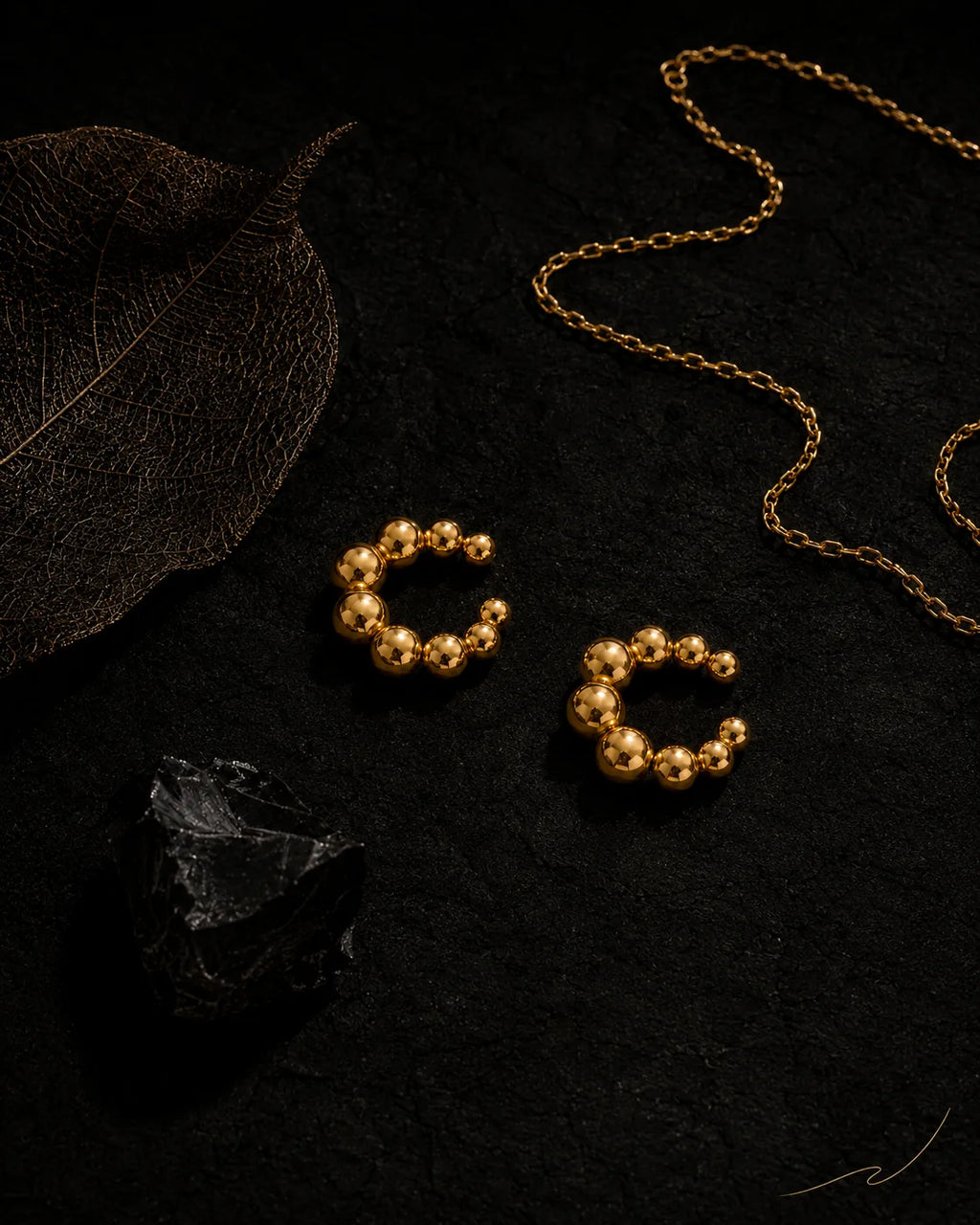 Gold Orbit ear cuff pair on black stone with dried leaf and gold chain, editorial dark styling