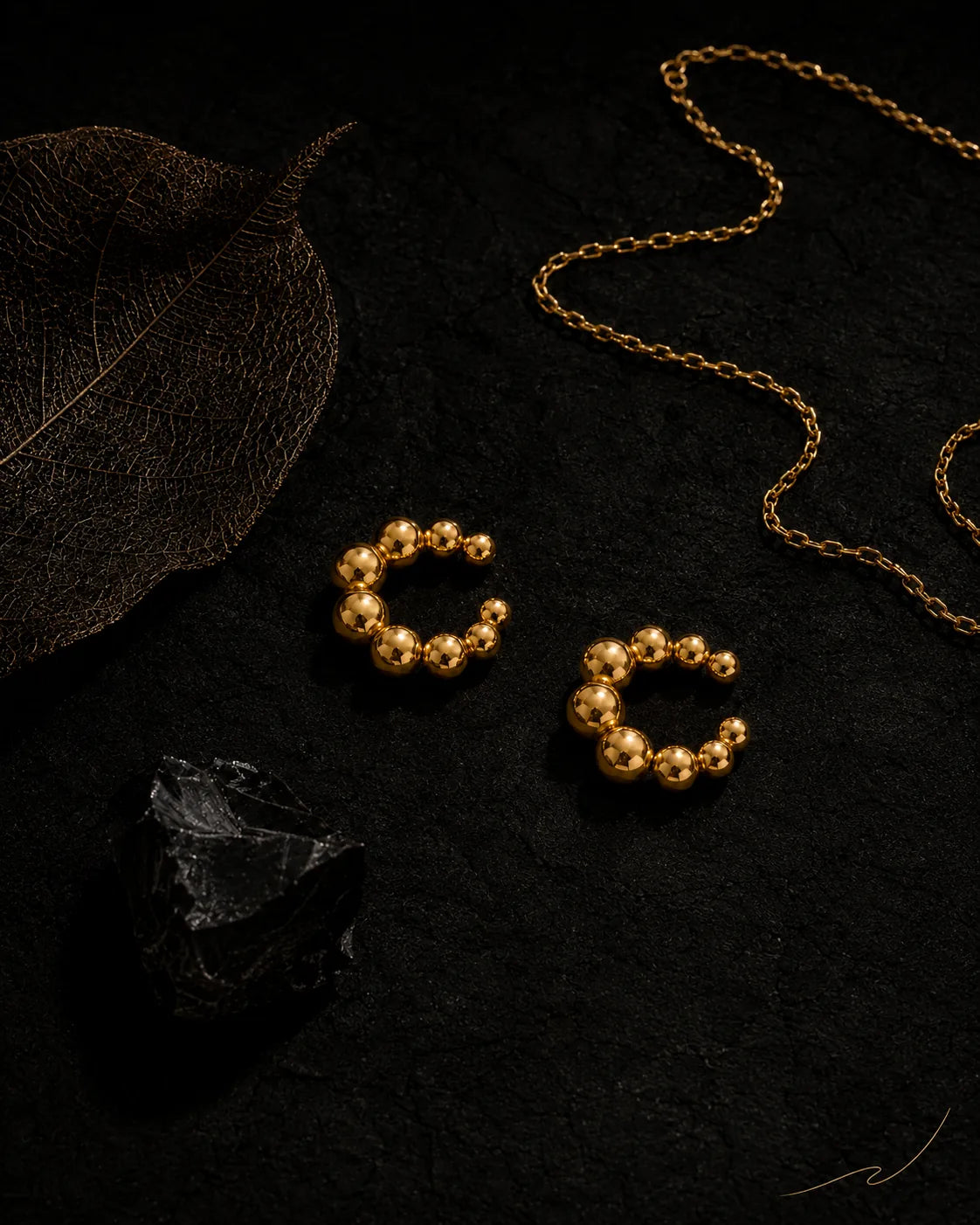 Gold Orbit ear cuff pair on black stone with dried leaf and gold chain, editorial dark styling