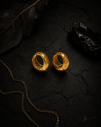 Gold Vault Huggie pair on cracked black stone with dried leaf and gold chain, editorial styling