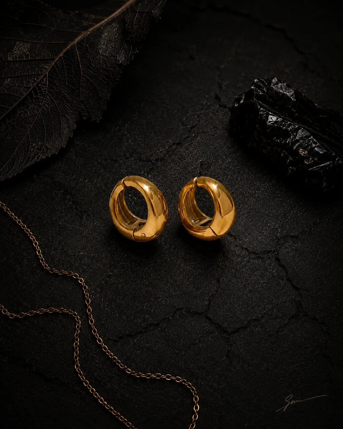 Gold Vault Huggie pair on cracked black stone with dried leaf and gold chain, editorial styling